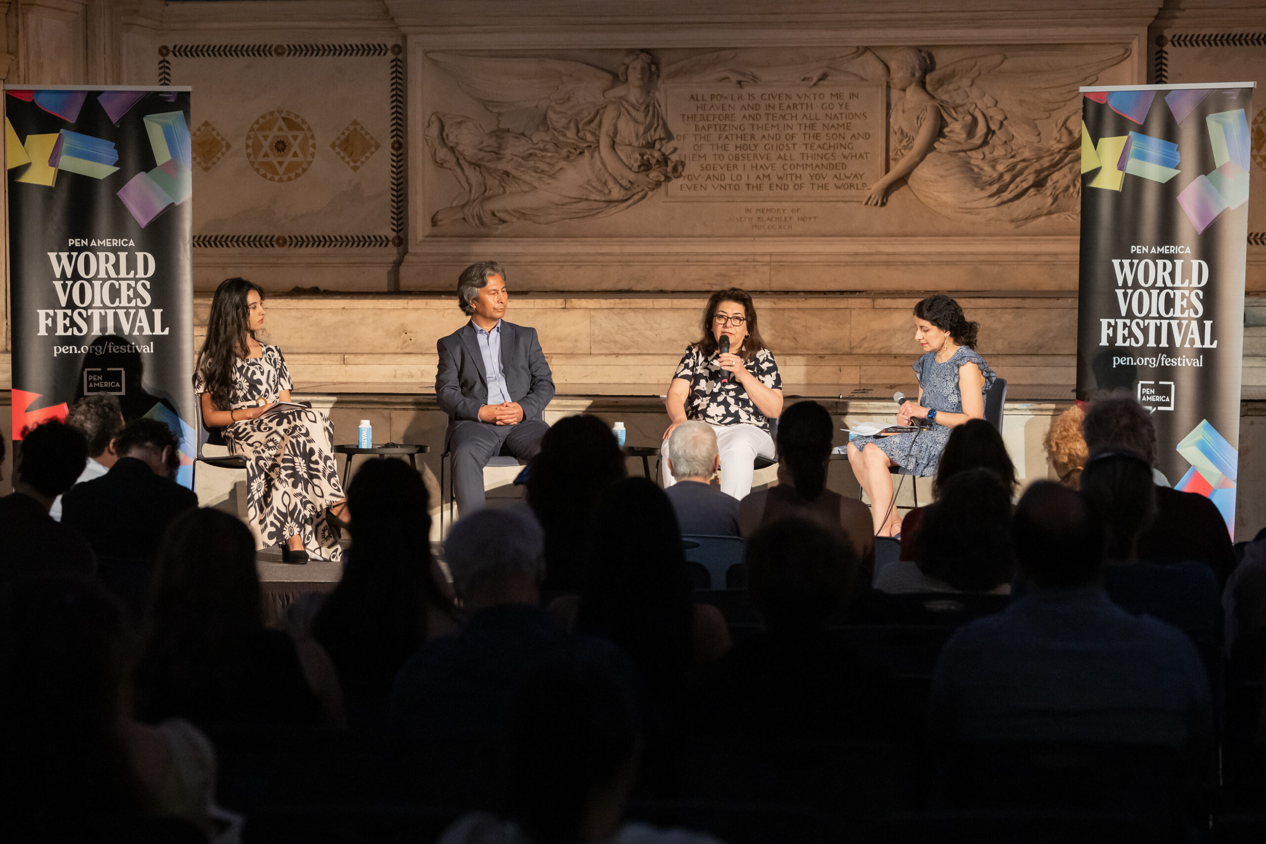 Four panelists sit on stage in front of an audience at the PEN America World Voices Festival. Two women and one man are seated, and one woman speaks into a microphone. Banners with the festival name flank the stage.