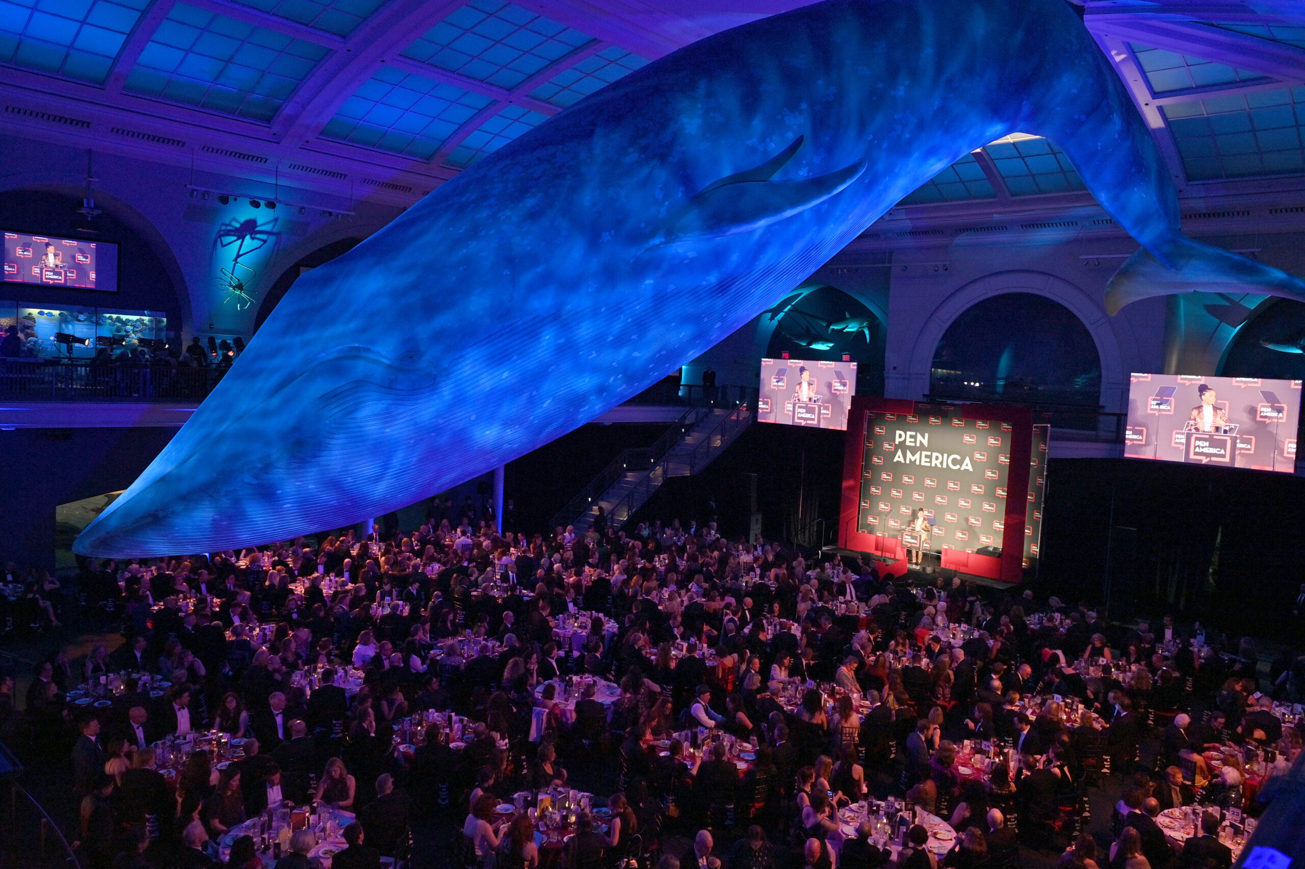 A large crowd attends a PEN America event in a grand hall under a life-sized blue whale model suspended from the ceiling, with stage, screens, and tables set for dinner.