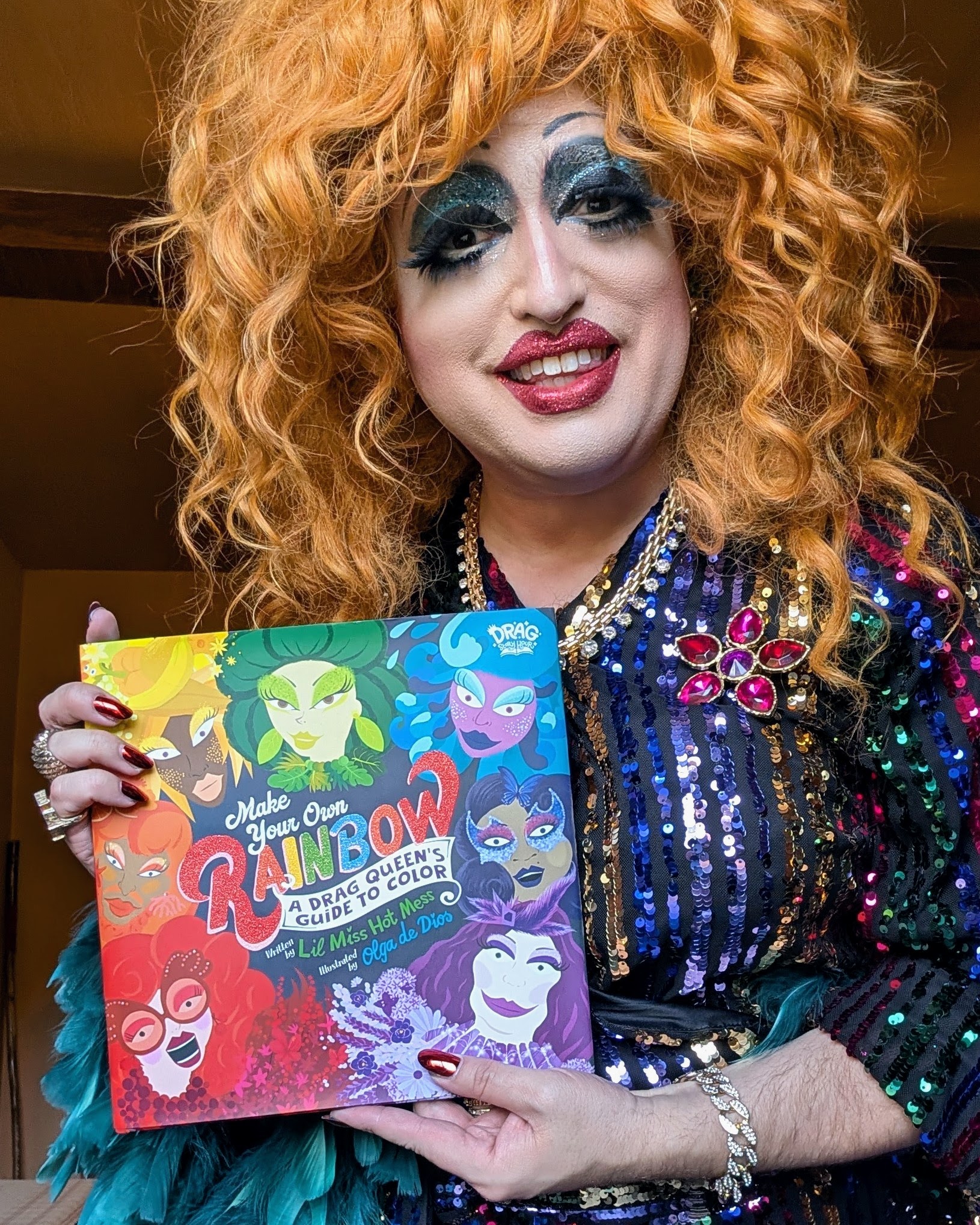 A person in colorful drag makeup and a vibrant orange wig smiles while holding a brightly illustrated “Make Your Own Rainbow: Drag Queen Guide & Coloring Book.” They wear a sequined outfit adorned with jeweled decorations.