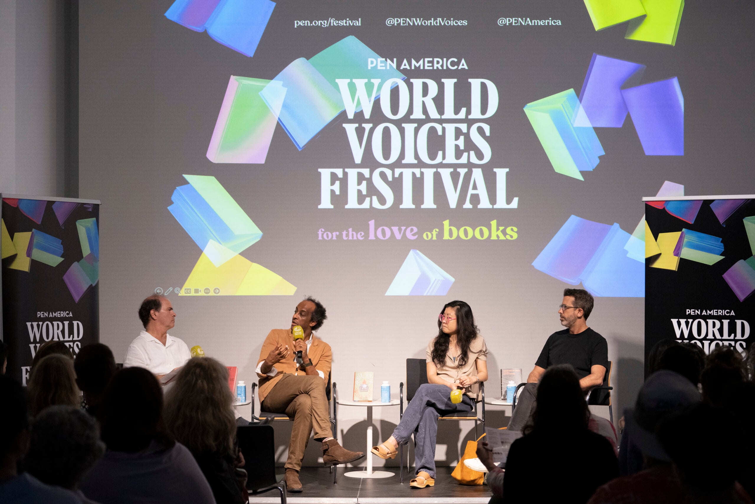 Four panelists sit on stage with microphones at the PEN America World Voices Festival. The backdrop reads “for the love of books” amid colorful graphics. Audience members are visible in the foreground.