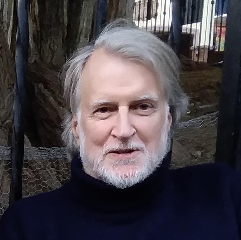 A man with light gray hair and a beard, wearing a dark turtleneck sweater, sits outdoors in front of a tree and a metal fence, smiling slightly at the camera.