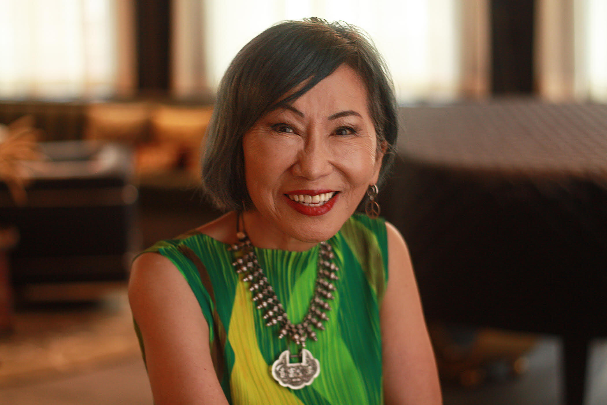 A smiling woman with short dark hair and a green patterned dress sits indoors, wearing a large statement necklace. Warm light fills the background, which is softly blurred.