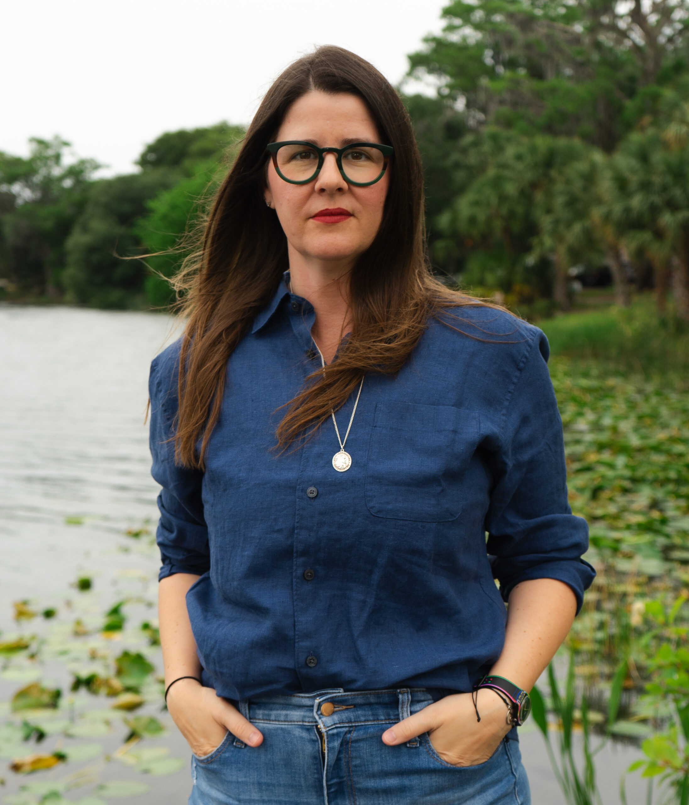 A woman with long brown hair and glasses stands by a lake with lily pads, wearing a blue button-up shirt and jeans, hands in her pockets, with trees in the background.