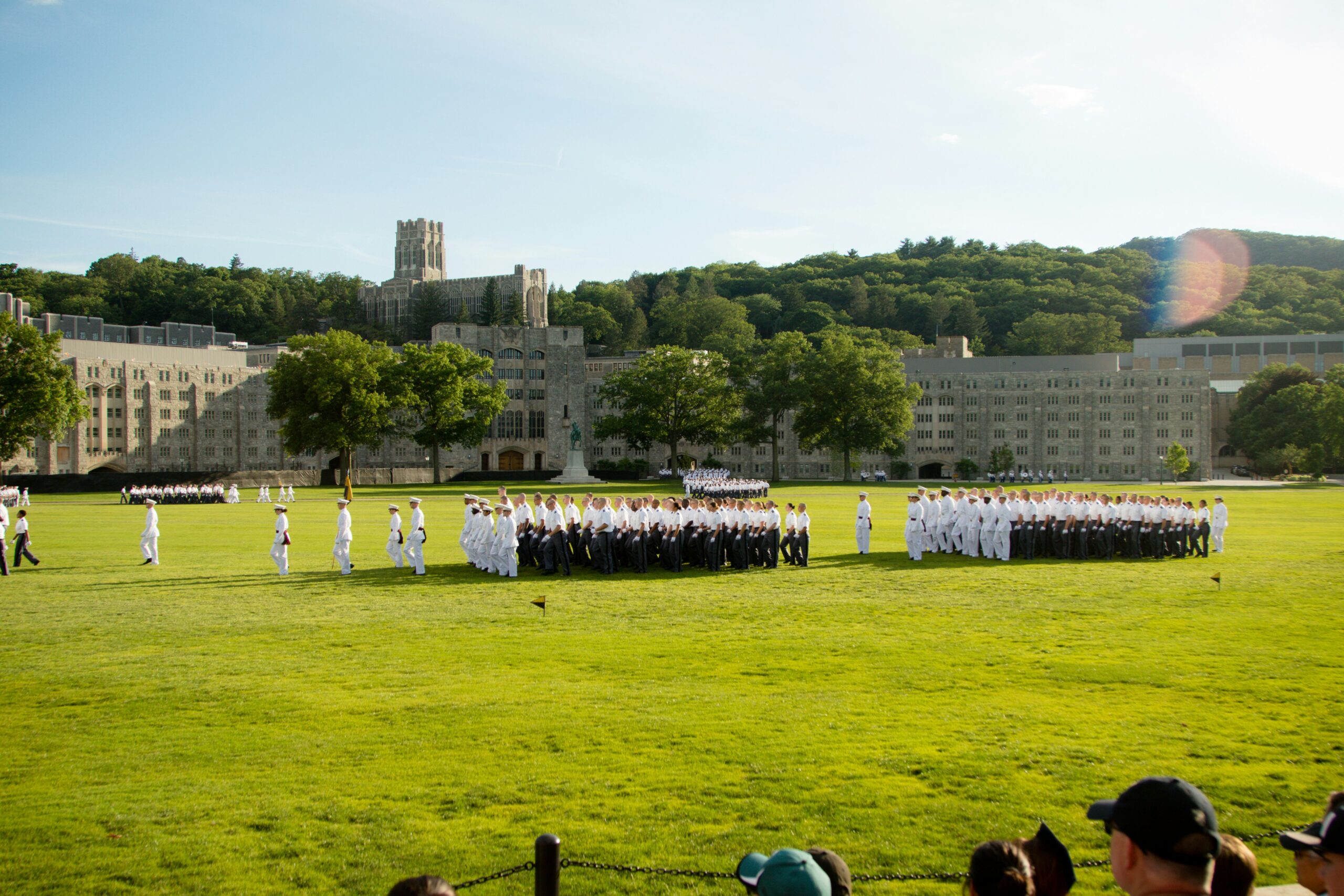 A group of uniformed cadets gathers on a large grassy field with a historic stone building and green hills in the background under a clear sky. Spectators are visible in the foreground.