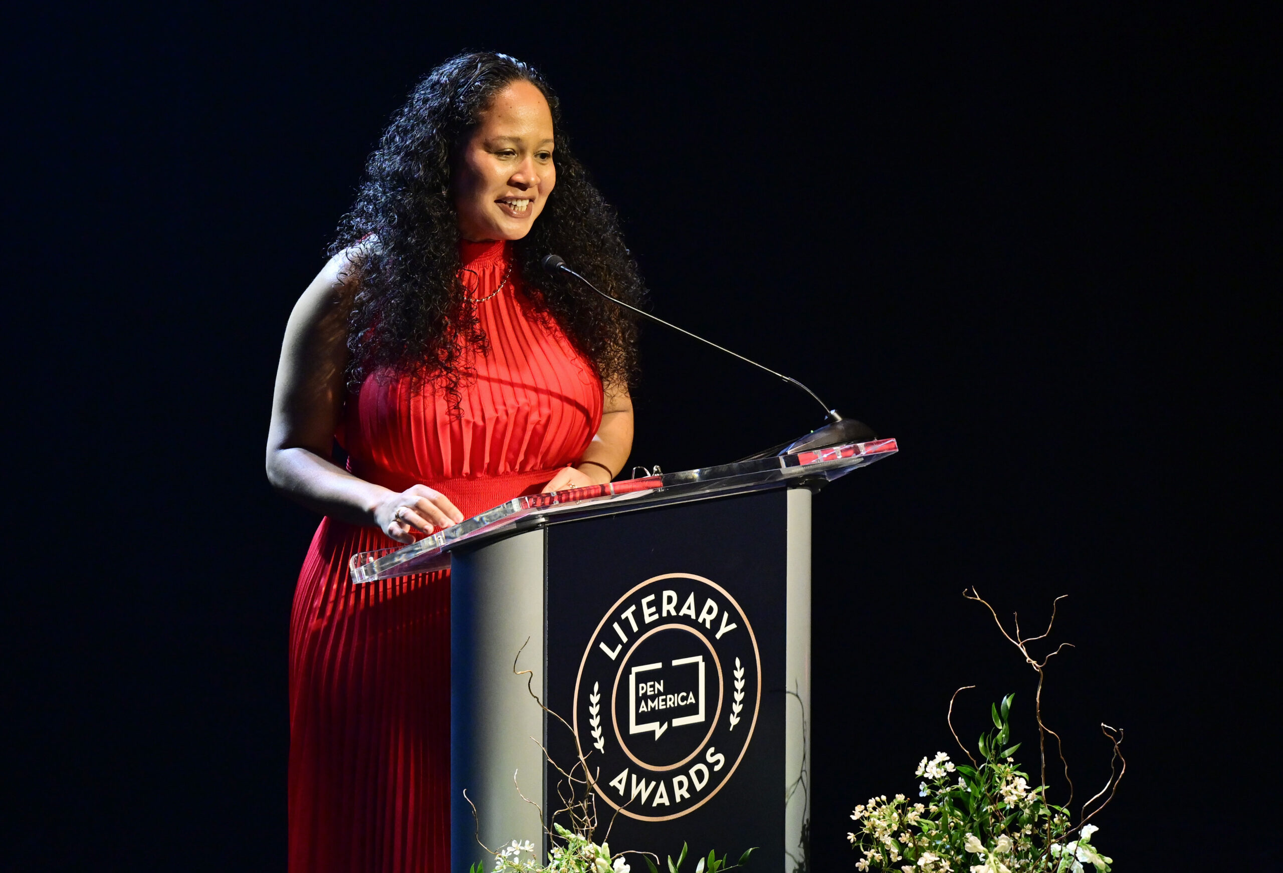 A woman in a red dress speaks at a podium labeled PEN America Literary Awards on a stage decorated with flowers.