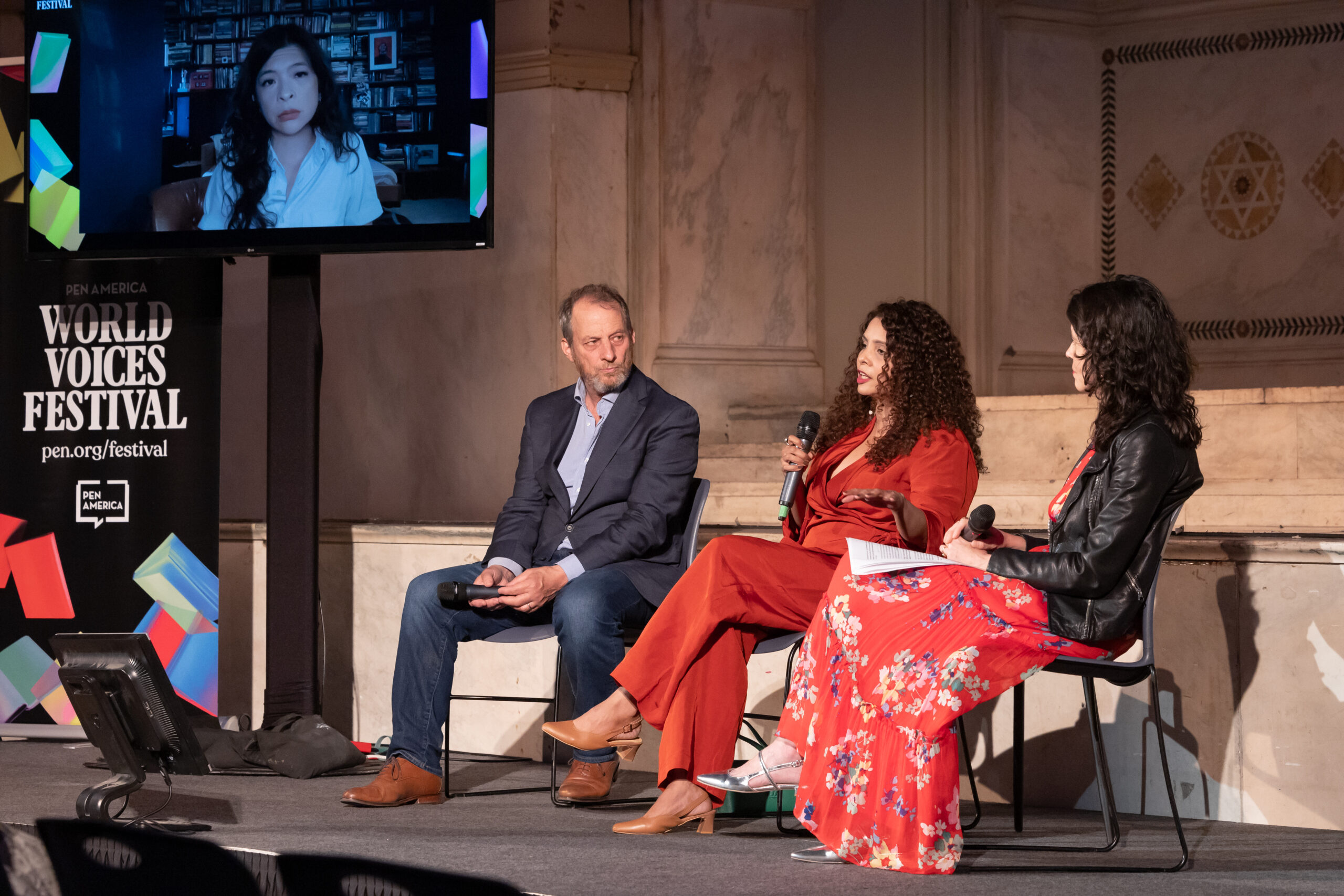 Three people sit on stage having a discussion; two women and one man face an audience, while a fourth woman appears on a large screen behind them. A sign reads PEN America World Voices Festival.