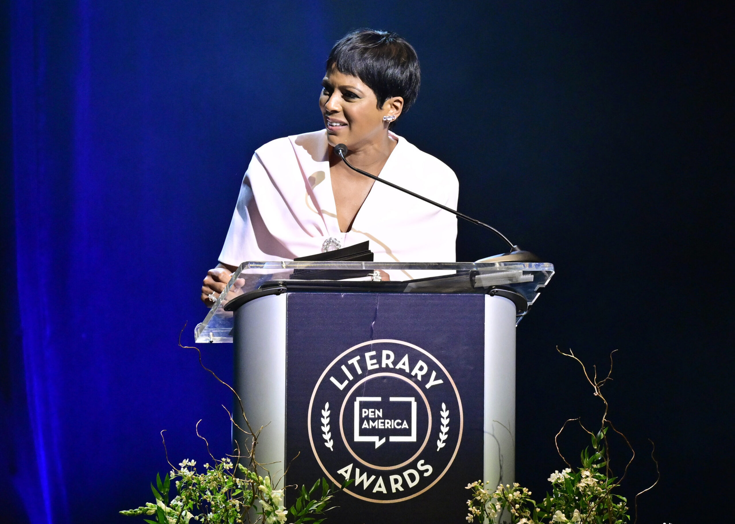 A woman stands at a clear podium with a PEN America Literary Awards sign, speaking into a microphone. She wears a white top and is surrounded by floral decorations, with a dark blue background behind her.