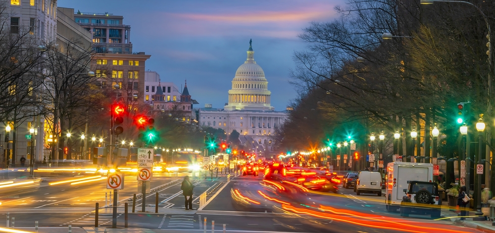 The U.S. Capitol building is illuminated at dusk, viewed from a busy city street with blurred car lights, traffic signals, and buildings lining both sides.