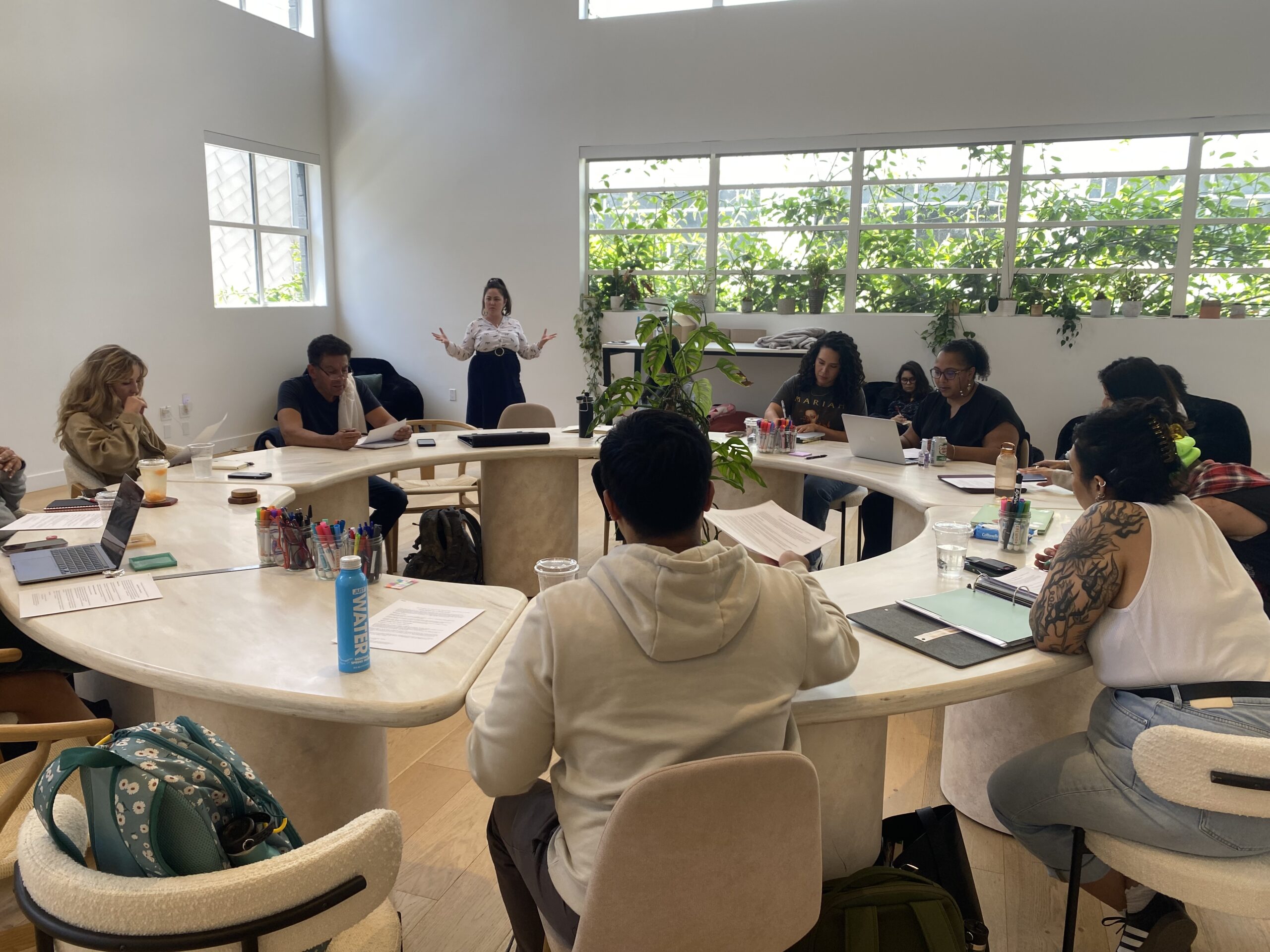A group of people sit around a large, U-shaped table in a bright room, actively listening to a woman standing and speaking at the front, with laptops, papers, and drinks on the table.