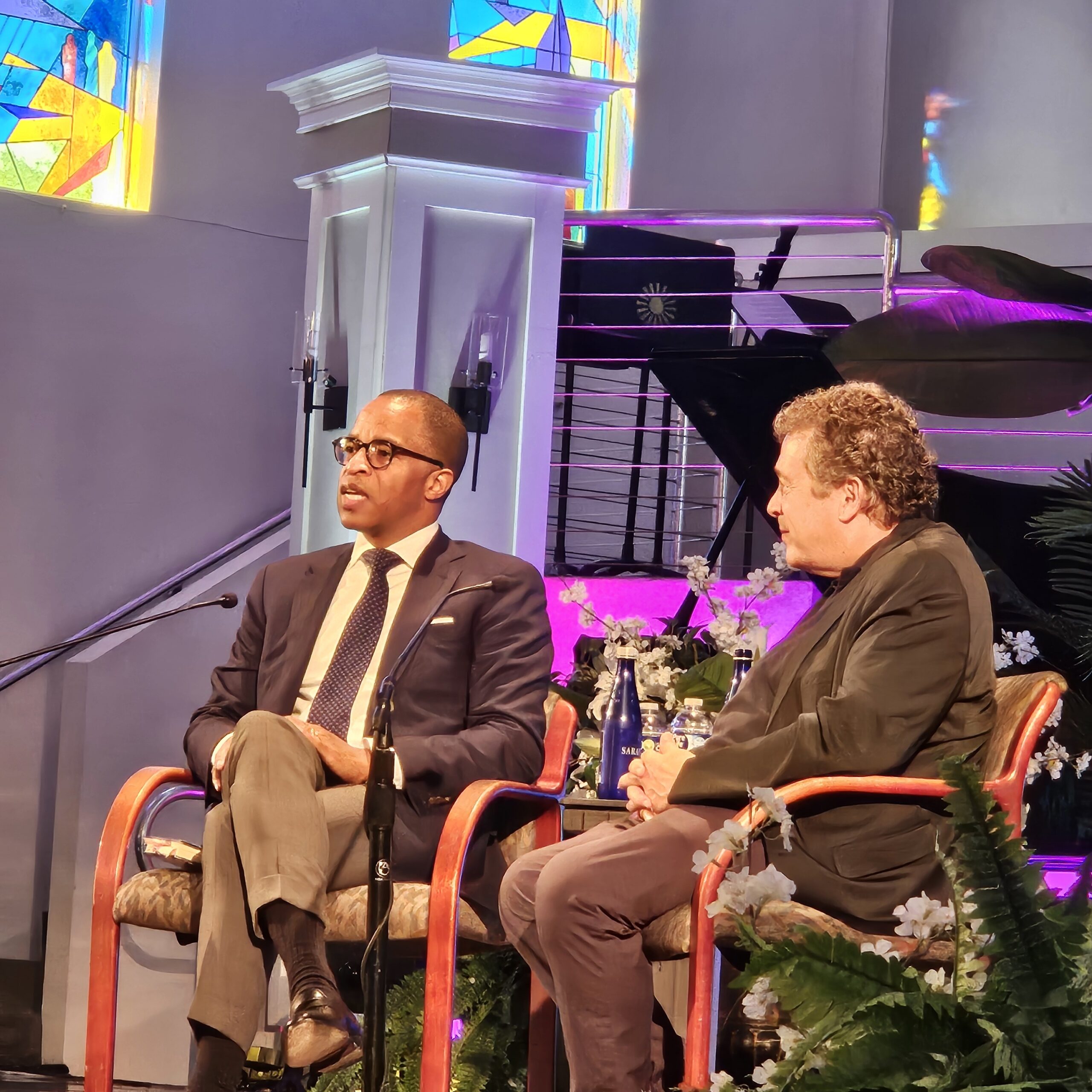 Two men sit on stage having a conversation in front of a piano, with microphones clipped to their shirts. Stained glass windows and flowers are visible in the background, suggesting a church or formal event setting.
