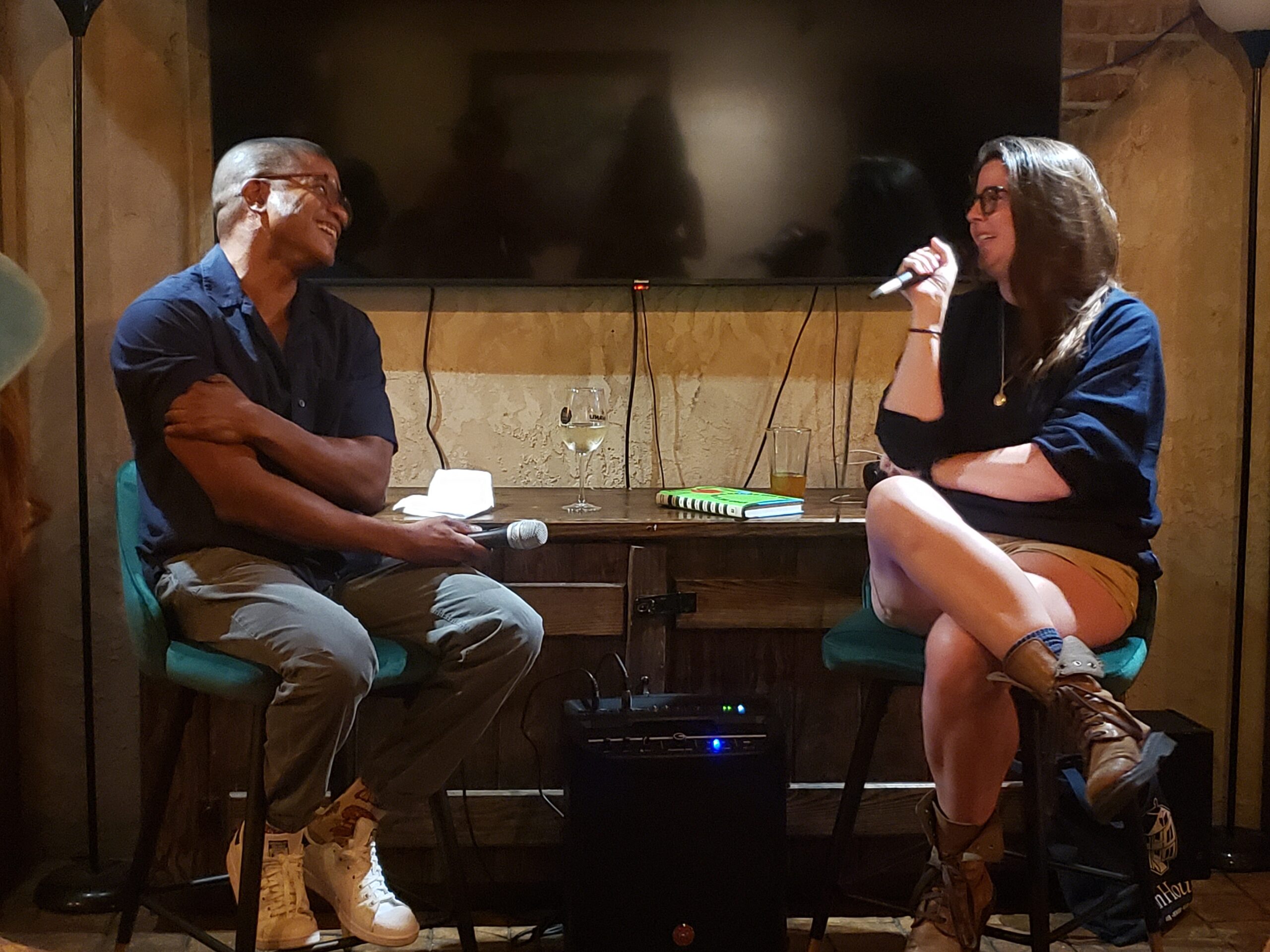 A man and a woman sit on stools, smiling and holding microphones during a conversation in a cozy room. A table with drinks and books is between them, and a TV is on the wall behind them.