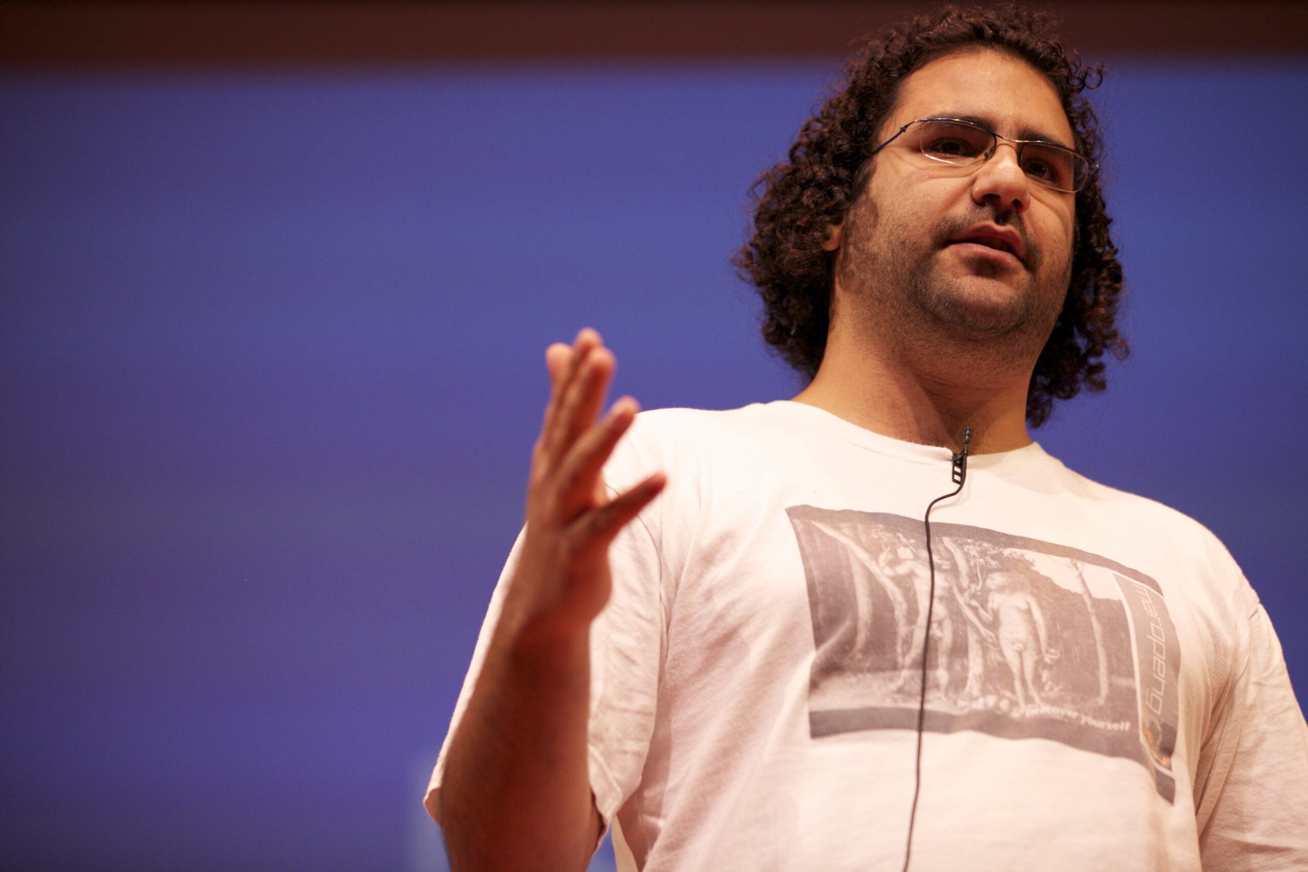 A man with curly hair and glasses speaks on stage, wearing a white t-shirt with a printed image and a headset microphone, gesturing with his hand against a plain, blurred background.