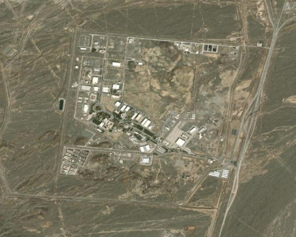 Aerial view of a large, fenced industrial complex with multiple buildings and roads, surrounded by barren, rocky terrain and intersected by a railway line.