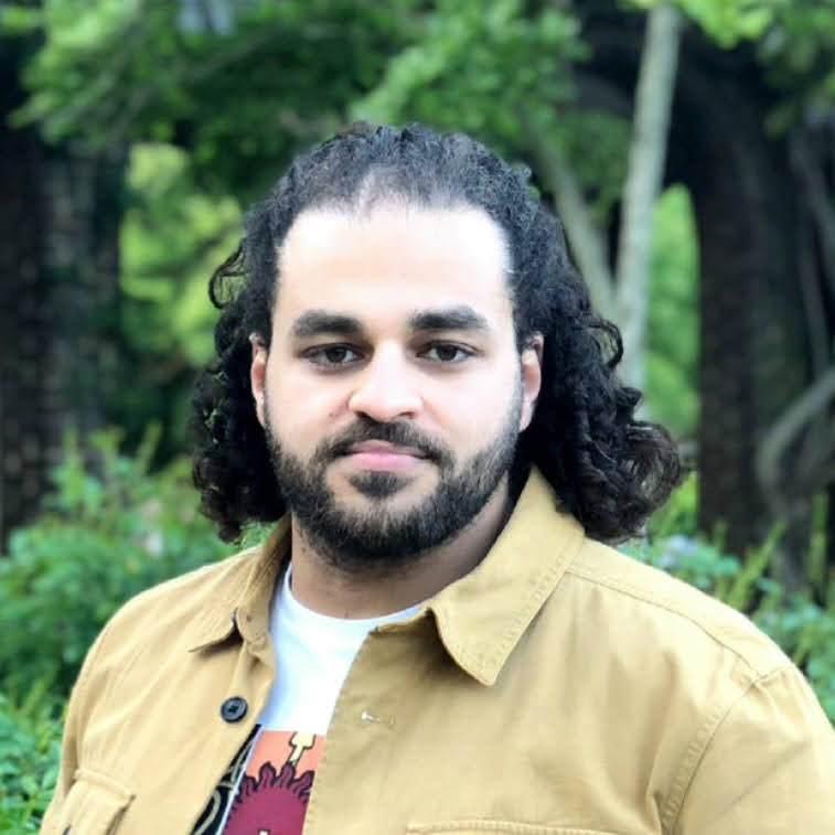A man with curly hair and a beard stands outdoors, wearing a tan jacket over a white shirt. Green foliage and trees are visible in the blurred background.