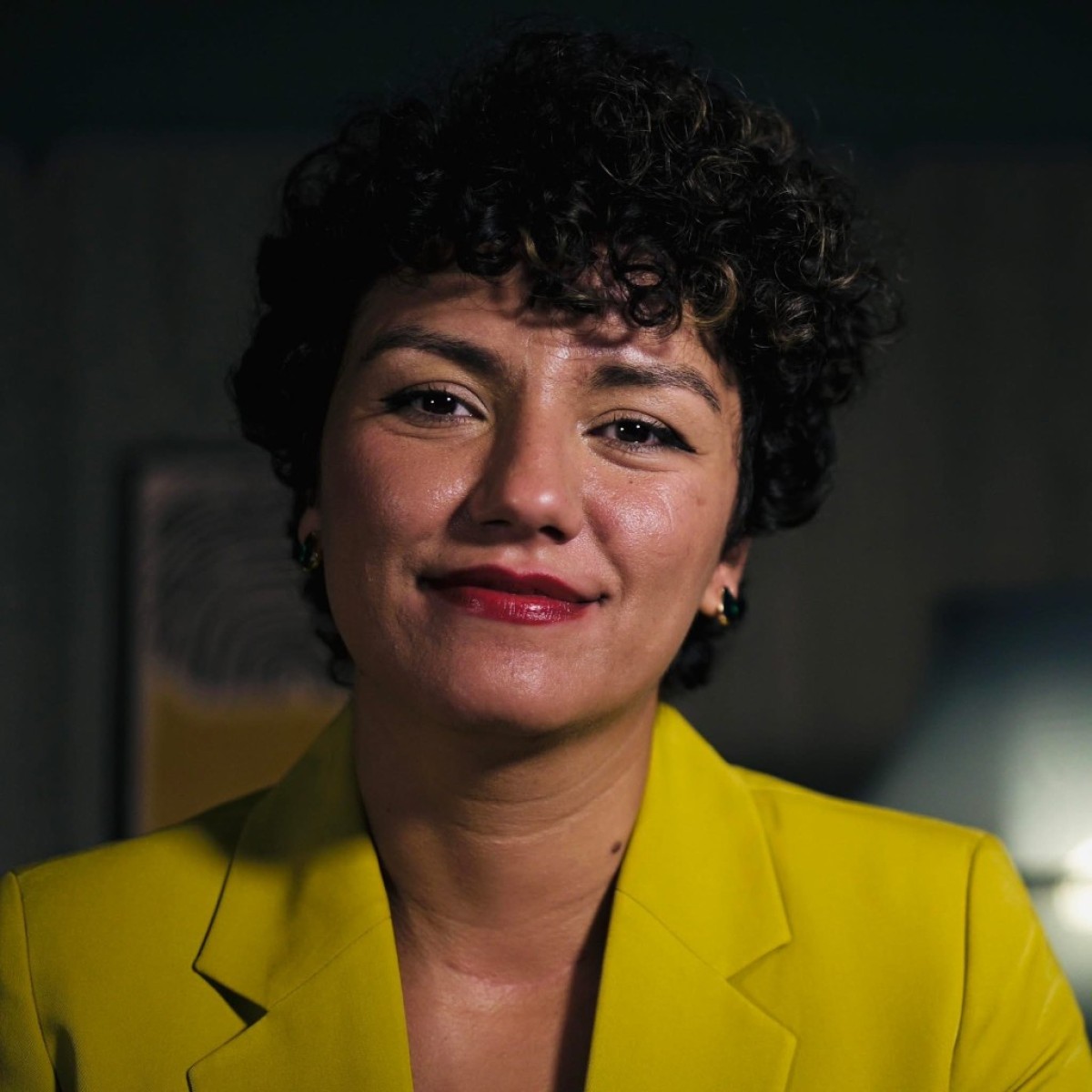A person with short curly hair, wearing a bright yellow blazer and red lipstick, smiles softly at the camera. The background is dimly lit with blurred objects and a lamp.