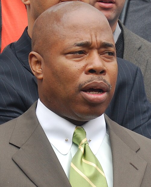 A man in a suit and striped green tie stands outside, looking ahead with a serious expression, mouth slightly open. Other people in business attire are partially visible in the background.