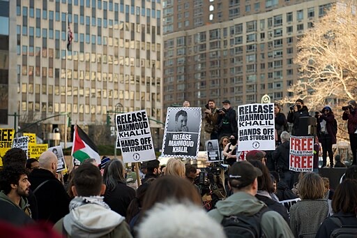 A crowd of protesters in a city holds signs reading “Release Mahmoud Khalil” and “Hands off our students,” with some waving a Palestinian flag. Photographers and speakers stand on a platform in the background.