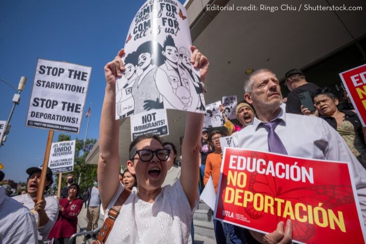 A diverse group of protesters hold signs in English and Spanish advocating for immigrant rights and against deportation. One sign reads EDUCACIÓN NO DEPORTACIÓN. The crowd looks passionate and engaged.