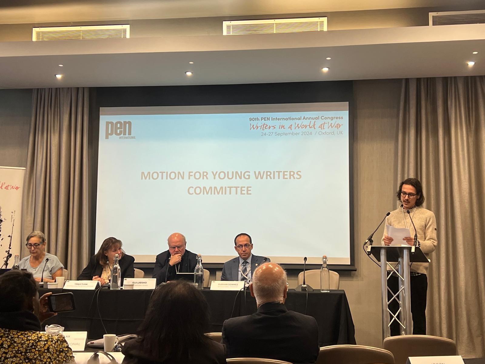 A person stands at a podium reading from a paper, while four people sit at a table on stage. Behind them, a screen displays MOTION FOR YOUNG WRITERS COMMITTEE at a PEN International Congress event.