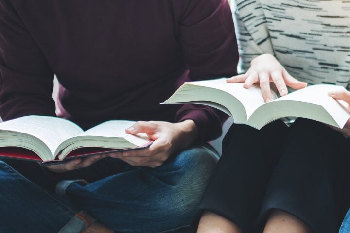 Two people sitting close together, each holding and reading a large open book. The focus is on their hands and books, with only their torsos and laps visible.