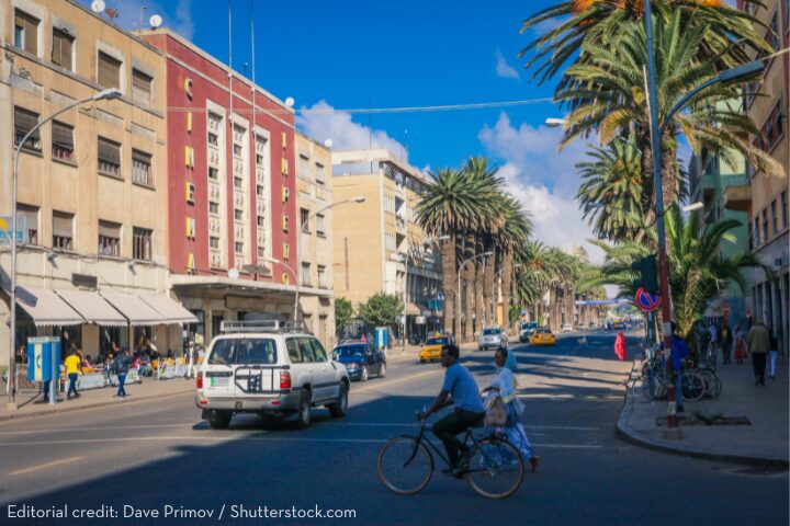 A busy street scene in Asmara, Eritrea, features people walking and cycling, cars driving by, and the historic Cinema Impero building with its art deco facade and large sign visible under a bright blue sky.