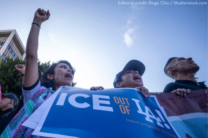 A group of people at a protest hold a large sign that reads ICE OUT OF LA! while raising their fists and shouting. Trees and buildings are visible in the background.