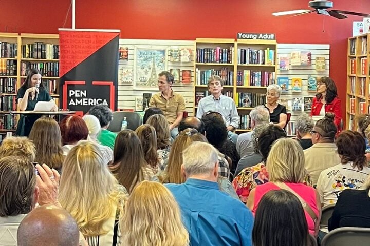 A woman speaks at a podium beside a Pen America banner, while four panelists sit on a stage facing an audience in a bookstore with shelves of books and a Young Adult section sign visible.