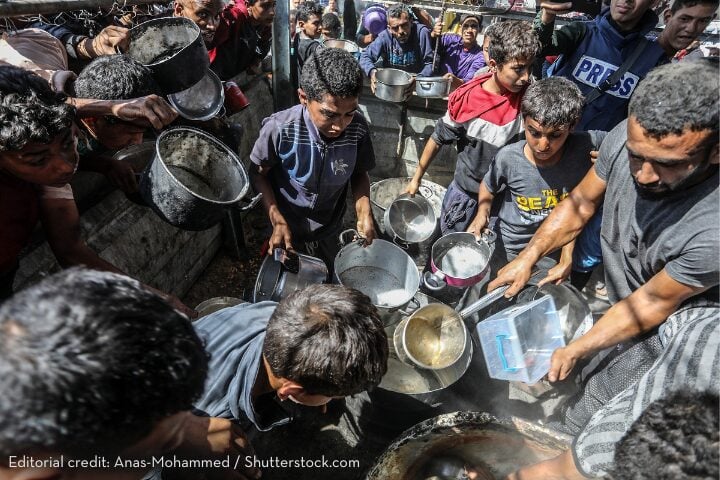 A group of people crowd around with empty pots and containers, reaching out in desperation, likely waiting for food or water distribution in a crisis setting. The scene is chaotic and urgent.