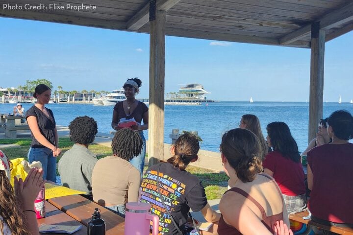 A group of people sits at picnic tables under a pavilion by the water, listening to two speakers standing in front of them. Sailboats and a modern building are visible in the background under a clear blue sky.