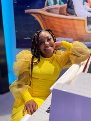 A smiling woman in a bright yellow dress with sheer sleeves sits at a white piano, resting her head on her hand. The background features part of a large photo or mural of people in a boat.