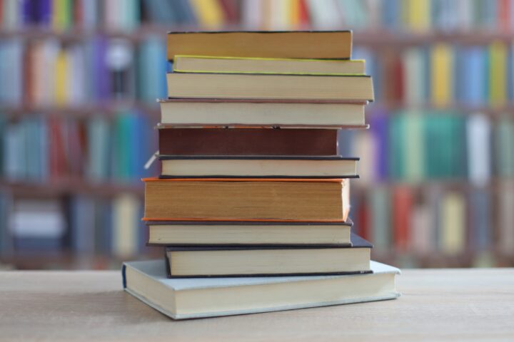 A stack of books is arranged on a wooden table with a blurred background of bookshelves filled with colorful books.