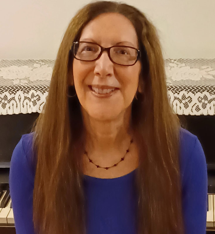 A woman with long brown hair and glasses smiles at the camera. She is wearing a blue top and a beaded necklace, sitting in front of a piano with a lace cover.