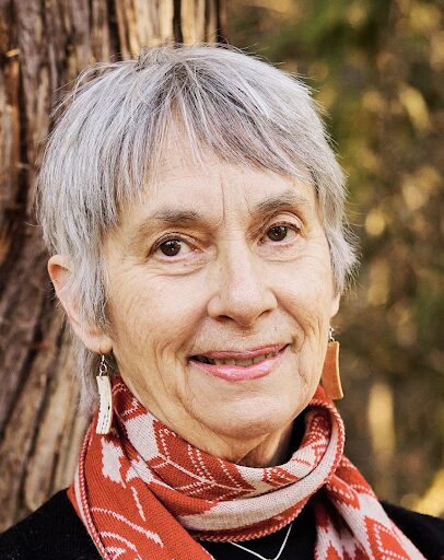 An older woman with short gray hair, wearing a red patterned scarf, wooden earrings, and a black top, smiles gently while standing outdoors in front of a tree trunk.