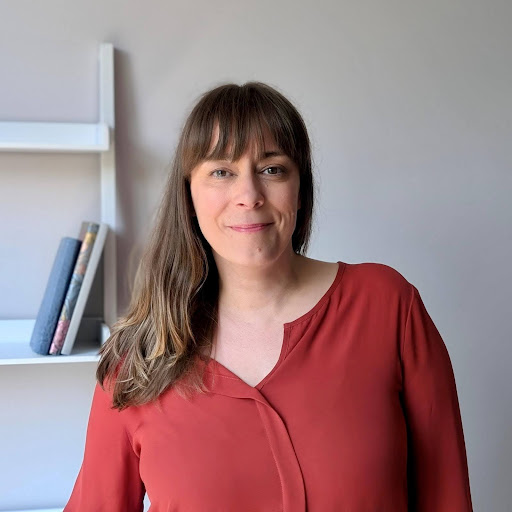A woman with long brown hair and bangs, wearing a rust-colored blouse, stands indoors in front of a gray wall with a white shelf holding several books. She is smiling gently at the camera.