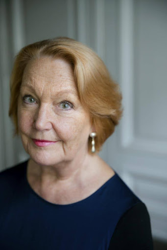 An older woman with short, light reddish-blonde hair and blue eyes, wearing a navy top and dangling earrings, stands in front of a softly lit, neutral background, looking slightly to the side and smiling gently.