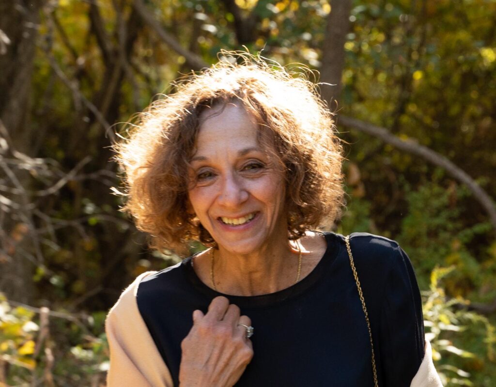 A woman with curly hair and a black top smiles outdoors in a sunlit forest, with trees and greenery in the background.