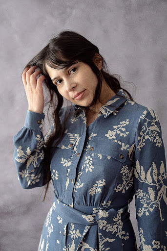 A woman with long dark hair wearing a blue dress with a light floral pattern stands against a soft gray background, gently touching her hair and smiling softly at the camera.