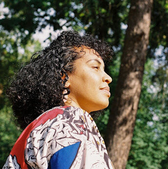A woman with curly hair stands outdoors in sunlight, looking thoughtfully into the distance. She is wearing a colorful patterned garment, and tall trees with green leaves fill the background.