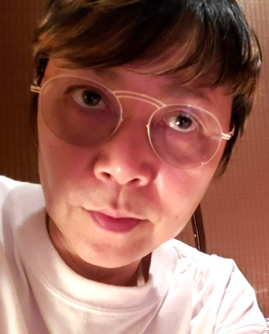 A person with short brown hair and round glasses is looking into the camera. They are wearing a white shirt and are indoors, with a warmly lit background.