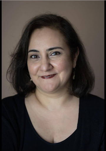 A woman with medium-length dark hair and light skin smiles gently at the camera. She is wearing a black top and subtle earrings, and is posed against a plain, neutral background.