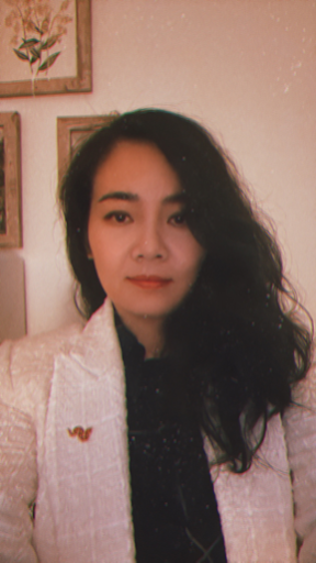 A woman with long, wavy black hair wearing a white textured blazer and black top, sitting indoors. Framed botanical artwork is visible on the wall behind her.
