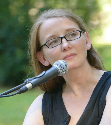 A woman with light skin and straight brown hair, wearing glasses and a sleeveless black top, sits outdoors in front of a microphone, looking thoughtfully to the side. The background is blurred greenery.
