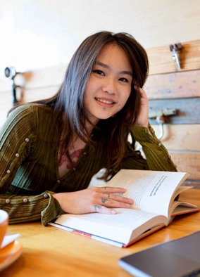 A young woman with long hair and facial piercings smiles while sitting at a wooden table, reading an open book. A laptop and coffee cup are nearby. She wears a green buttoned shirt.
