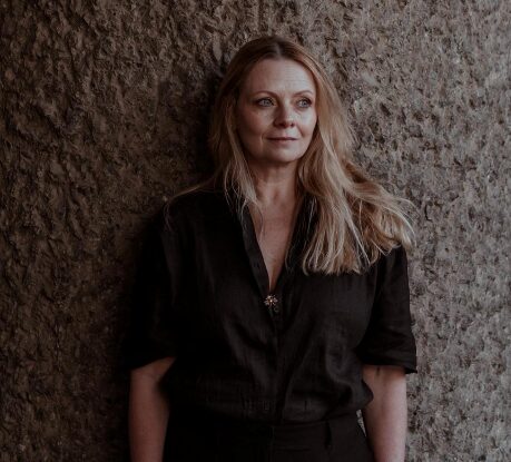 A woman with long blonde hair wearing a dark shirt stands against a textured stone wall, looking slightly to the side with a calm expression.