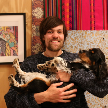 A smiling man with brown hair and a beard holds a black, brown, and white dog in his arms. Colorful patterned fabrics and framed artwork decorate the background.