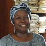 A woman wearing a patterned headwrap and earrings smiles in front of a shelf stacked with documents and papers.