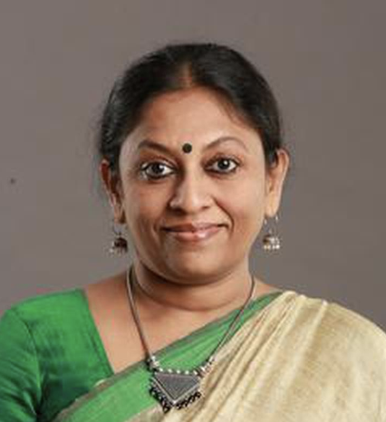 A woman with dark hair tied back, wearing a green and beige saree, silver jewelry, and a black bindi, smiles softly in front of a plain gray background.