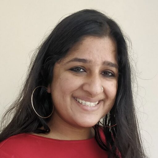A woman with long dark hair, wearing a red top and large hoop earrings, smiles at the camera in front of a plain light-colored background.