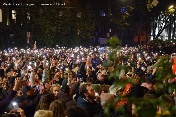 A large crowd gathers outdoors at night, many people holding up smartphones with flashlights on, creating a sea of lights. Some are waving flags, and trees are visible in the background.