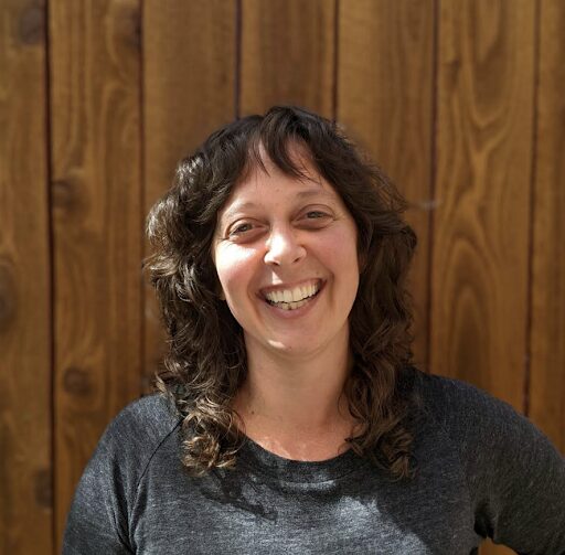 A woman with curly brown hair and a gray shirt smiles warmly at the camera while standing in front of a wooden fence in bright sunlight.